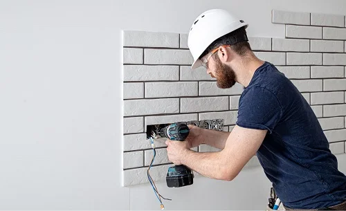 Electrician installing electrical outlets on a wall, wearing a hard hat and using a power drill, emphasizing professional electrical services in Newport Beach.